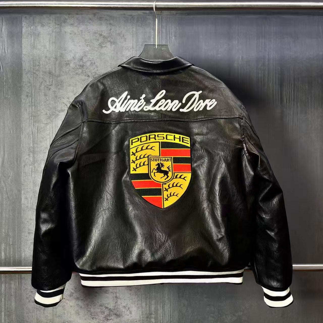 Black leather jacket with Porsche logo and 'Aime Leon Dore' text on a metallic surface.