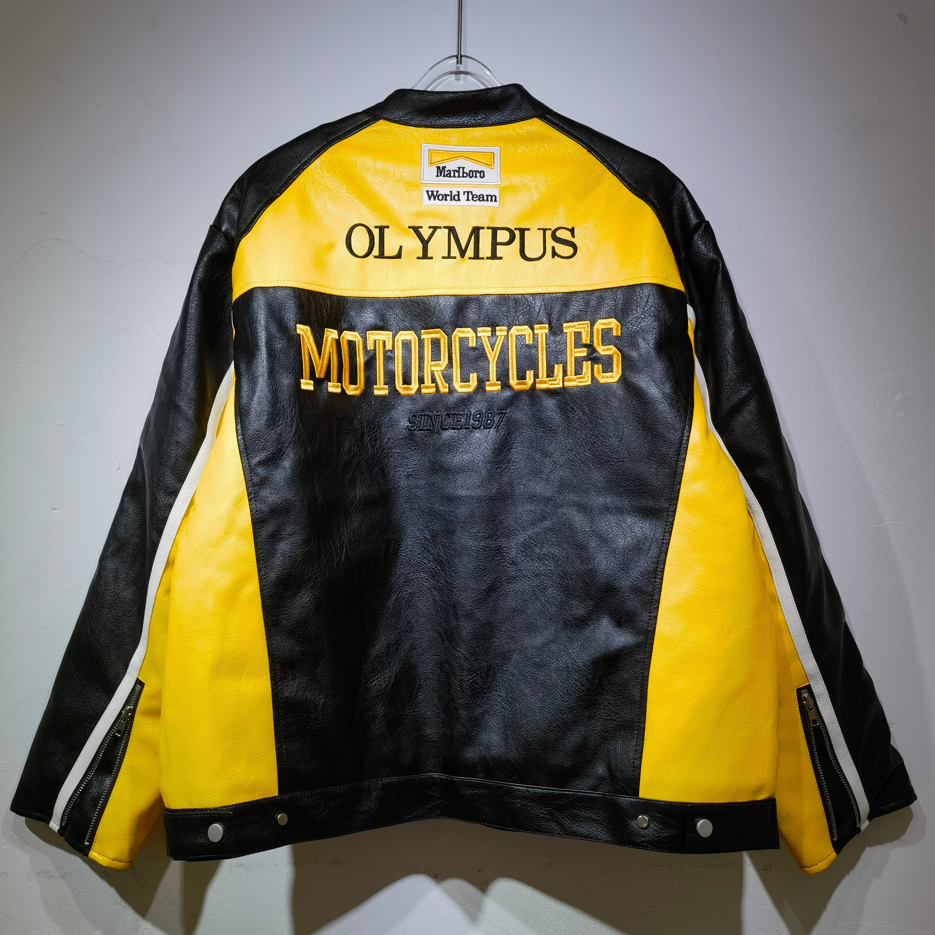 Black and yellow leather jacket with 'Olympus Motorcycles' branding on a gray background