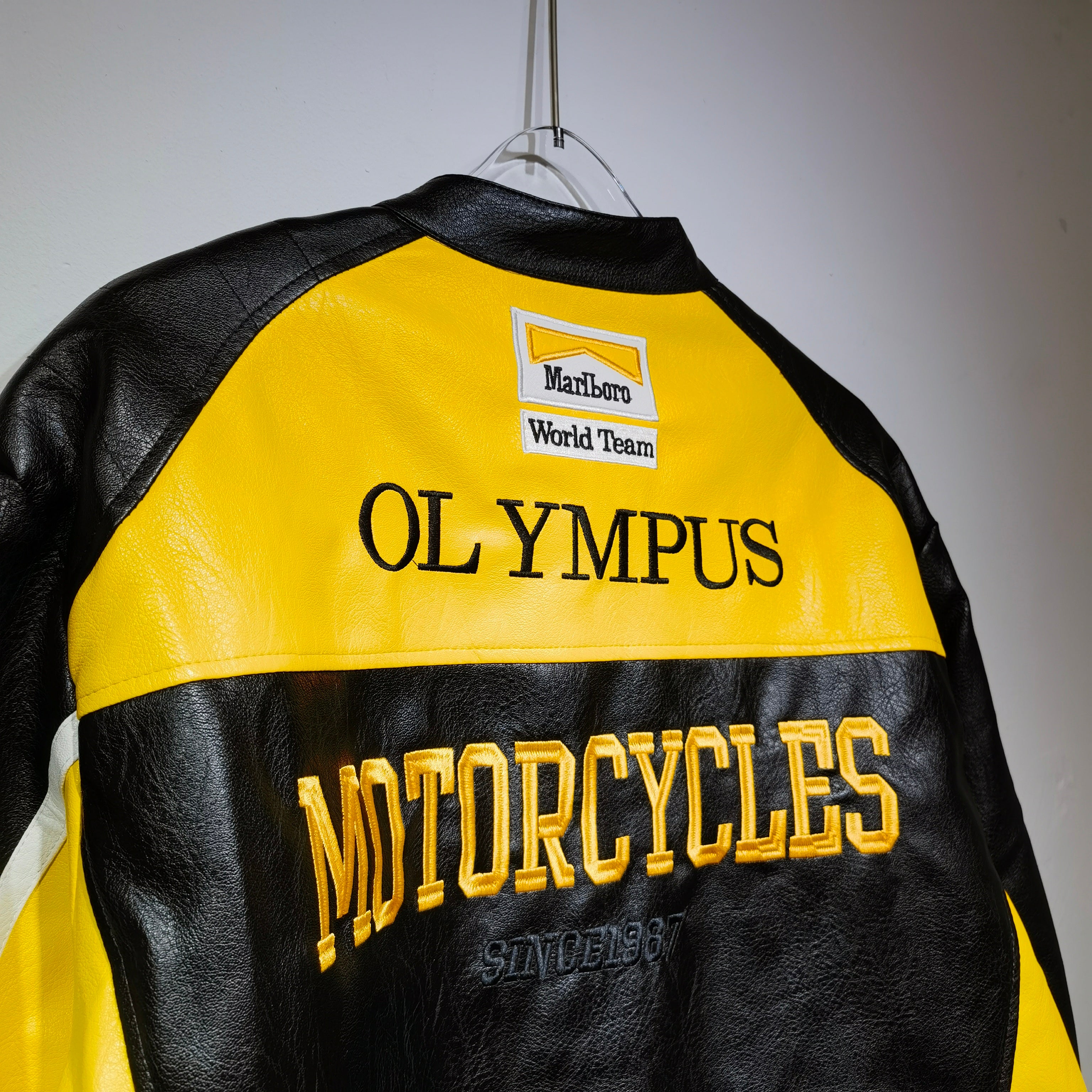 Black and yellow motorcycle jacket with 'Olympus Motorcycles' branding on a gray background